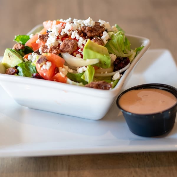 A fresh salad with lettuce, tomatoes, avocado, nuts, and feta cheese, with a side of dressing.