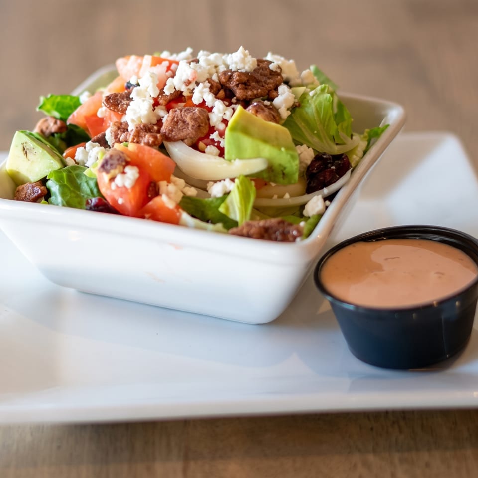 A fresh salad with lettuce, tomatoes, avocado, nuts, and feta cheese, with a side of dressing.