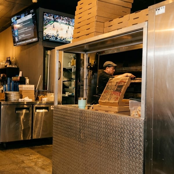 A chef in a hat places a pizza into a large oven behind a counter with stacked pizza boxes.