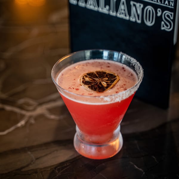 A vibrant pink cocktail with a sugar rim and dried orange slice, next to an Italiano's menu.