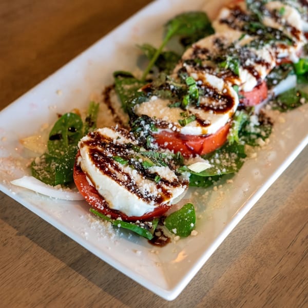Caprese salad with sliced mozzarella, tomatoes, spinach, balsamic glaze, and grated cheese on a white plate.
