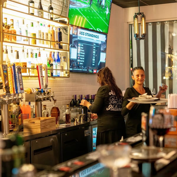 Two women work behind a well-stocked bar with beer taps, liquor bottles, and TVs showing sports.