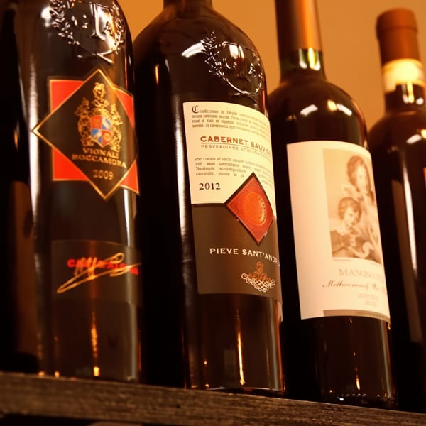 Several dark wine bottles with various labels, including "Vignali Roccamora" and "Cabernet Sauvignon," on a wooden shelf.