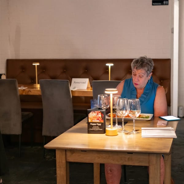 A woman in a blue top sits at a restaurant table with wine glasses and a plate of food.