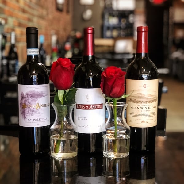 Three bottles of red wine with two red roses in small vases on a reflective surface.