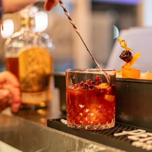 Bartender stirring a red cocktail with fruit garnish and orange slices on a bar.