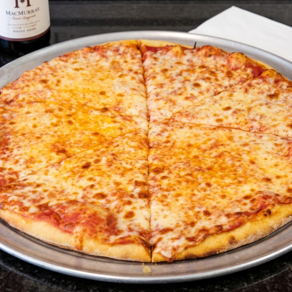 A large cheese pizza on a metal tray, with a bottle of wine and napkins in the background.