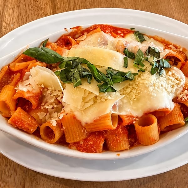 Baked rigatoni pasta with melted cheese, tomato sauce, and fresh basil in a white dish.