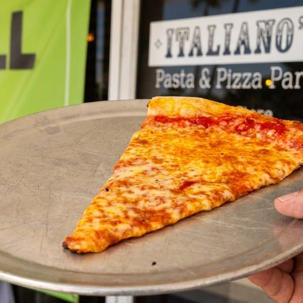 Hand holding a metal tray with a slice of cheese pizza in front of Italiano's Pizza Parlor.