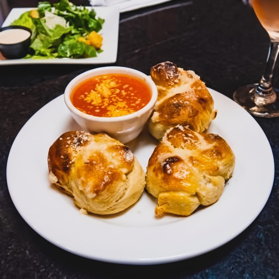 Three golden-brown garlic knots with parmesan cheese, a side of marinara, and a salad in the background.